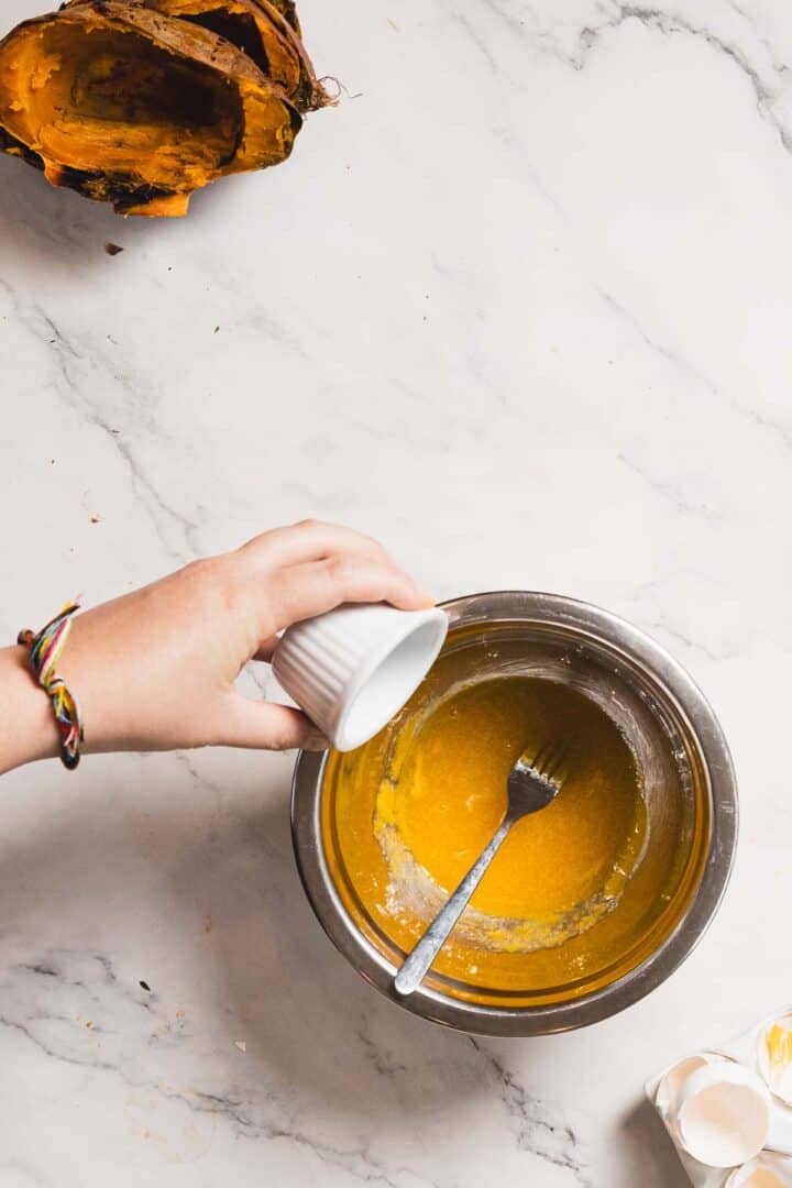 A hand pours a small white cup of liquid into a stainless steel bowl containing a yellow mixture, perfecting the recipe for Sweet Potato Crème Brûlée. A fork is placed inside the bowl. On the marble surface, there is a cut-open pumpkin with seeds removed and some scattered eggshells.