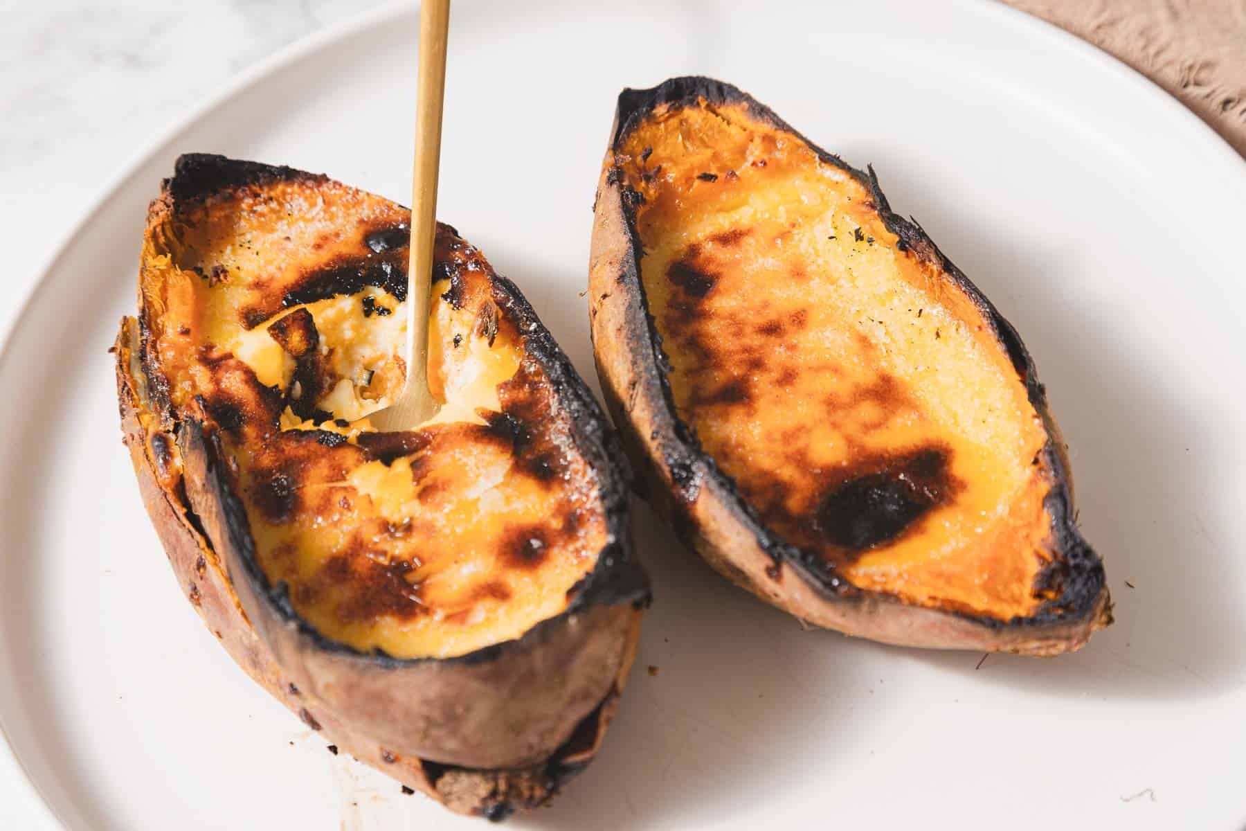 Two baked sweet potato halves with slightly charred, crème brûlée-like tops are on a white plate. A wooden spoon is scooping into one of the halves, revealing the soft interior of this delightful recipe.