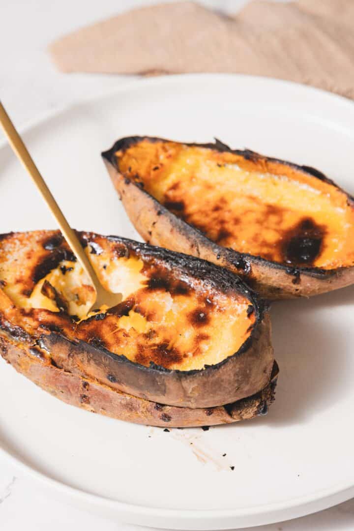 Two roasted sweet potato halves rest on a white plate, their edges slightly charred like a Sweet Potato Creme Brûlée. A gold spoon elegantly scoops the soft, orange flesh from one half. In the background, a beige cloth adds to the cozy setting of this delightful recipe scene.