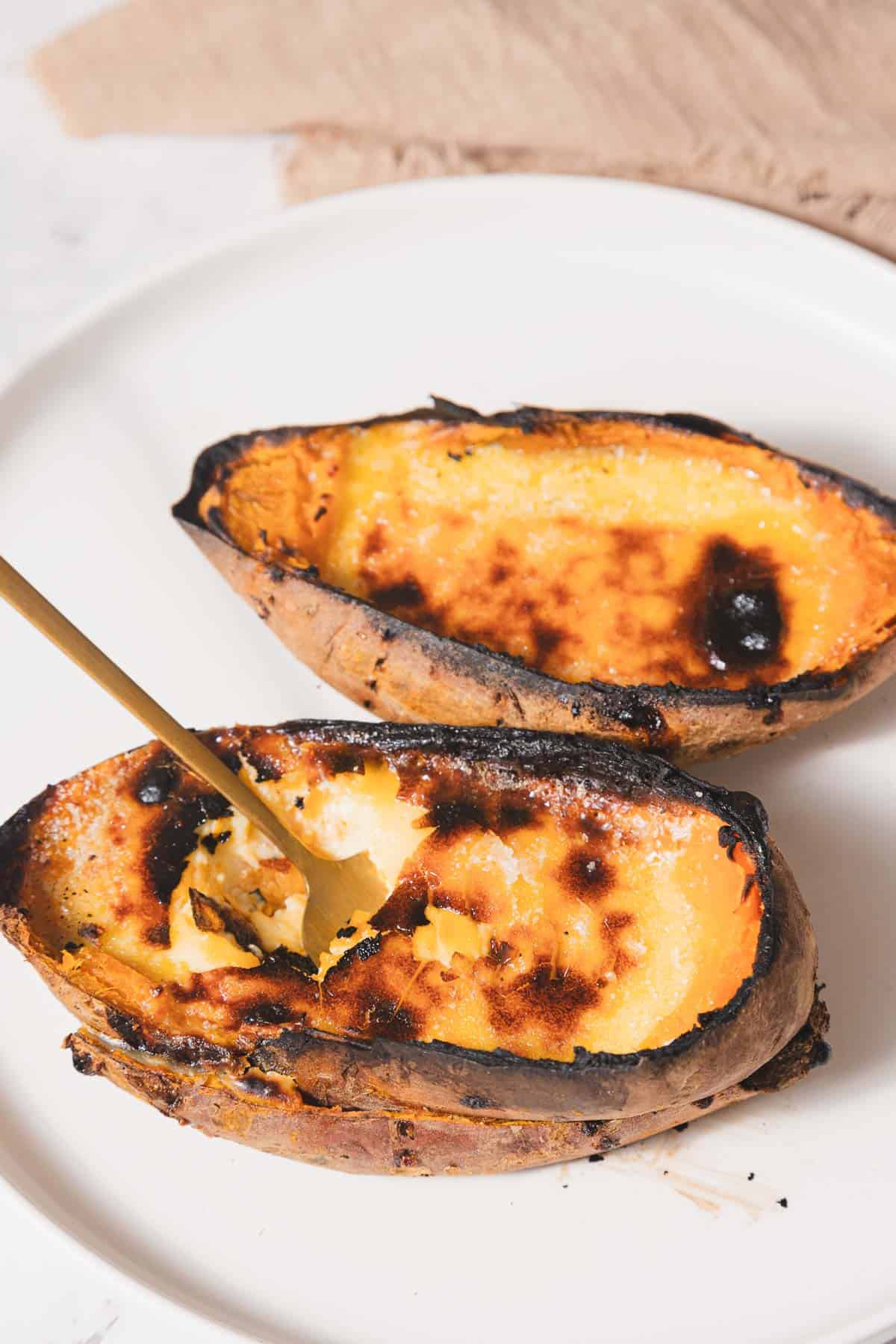 Two roasted sweet potato halves, reminiscent of creme brûlée with their slightly charred tops, rest on a white plate. A gold fork playfully pierces one half, ready for tasting. In the background, a beige cloth adds a touch of understated elegance to this deceptively simple yet delightful dish.