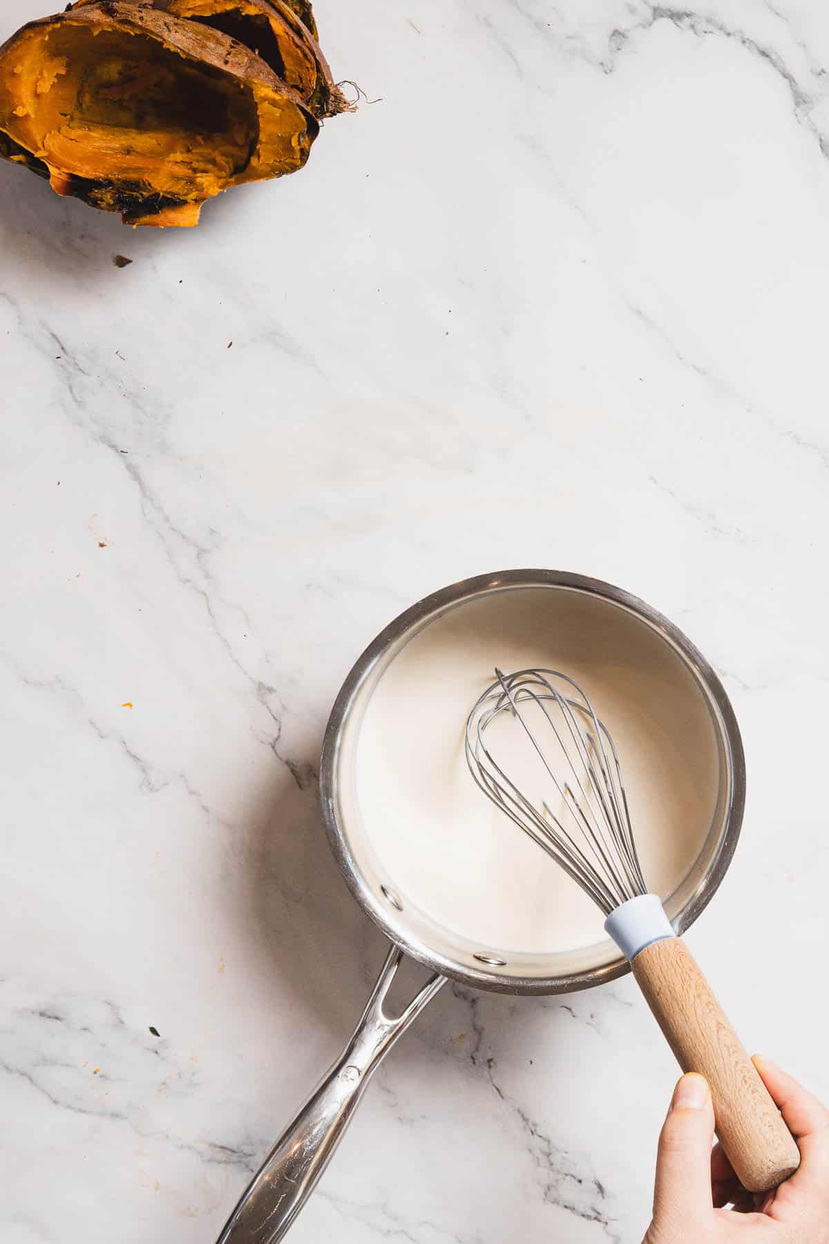 A hand expertly whisking a creamy mixture in a saucepan on a marble countertop, crafting the best recipe. A piece of pumpkin shell tempts in the upper left corner, hinting at the delightful flavors along with sweet potato.