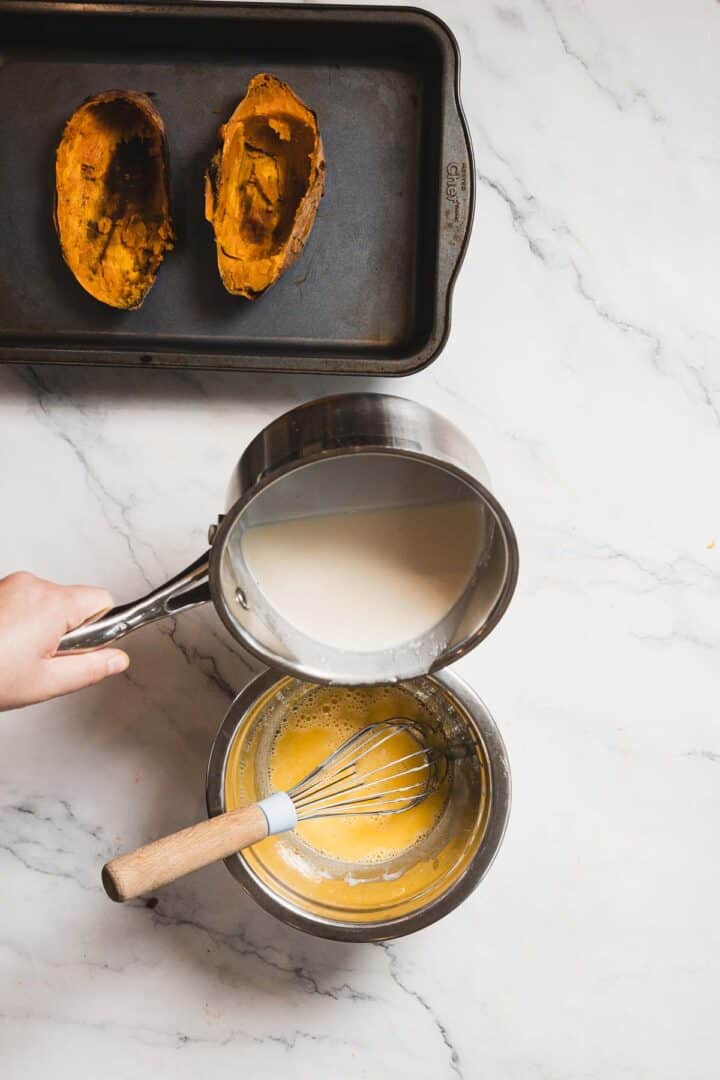 A person pours a white liquid from a saucepan into a bowl containing a mixture being whisked, perfecting a creamy creme brûlée recipe. Two empty roasted sweet potato halves rest on a baking tray against the elegant backdrop of the marble countertop.