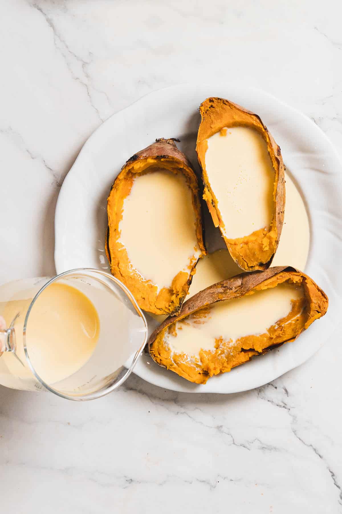 A white plate showcases three halved baked sweet potatoes brimming with a creamy sauce, a delightful twist on the classic Sweet Potato Creme Brûlée. To the left, on the marble surface, a glass pitcher holds more of this indulgent sauce.