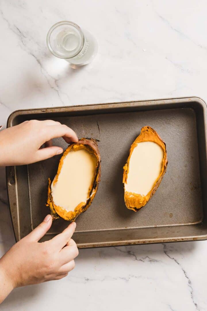 Two baked sweet potatoes on a baking sheet, each filled with a creamy mixture that rivals crème brûlée. A person's hands are adjusting one of the potatoes. A glass of water rests in the top left corner on a marble surface, complementing this indulgent recipe tableau.