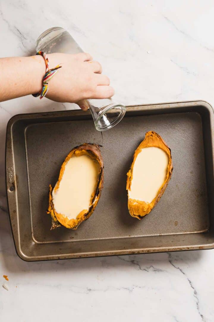 A baking tray with two hollowed sweet potato skins filled with a creamy recipe reminiscent of crème brûlée. A person's hand, adorned with a colorful bracelet, pours water from a glass into the tray. The surface is a light marble pattern.