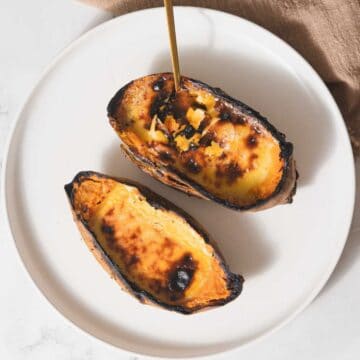 Two roasted sweet potato halves rest on a round white plate, with a gold-colored fork gently inserted into one half. The sweet potatoes have a charred, caramelized surface reminiscent of creme brûlée. A brown cloth is partially visible in the background, adding warmth to the scene.