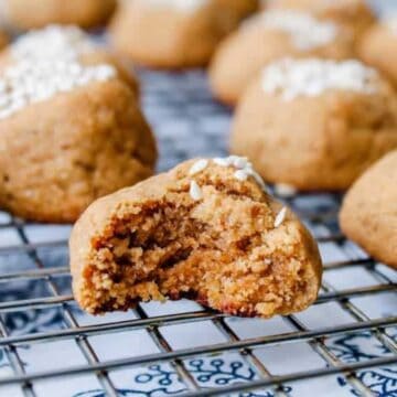 Cookies with a crumbly texture are cooling on a wire rack, tempting anyone to start stealing them straight from the jar. The cookies have a sprinkling of white sugar on top. One cookie in the foreground has a bite taken out, revealing its soft interior. The rack rests on a patterned surface.