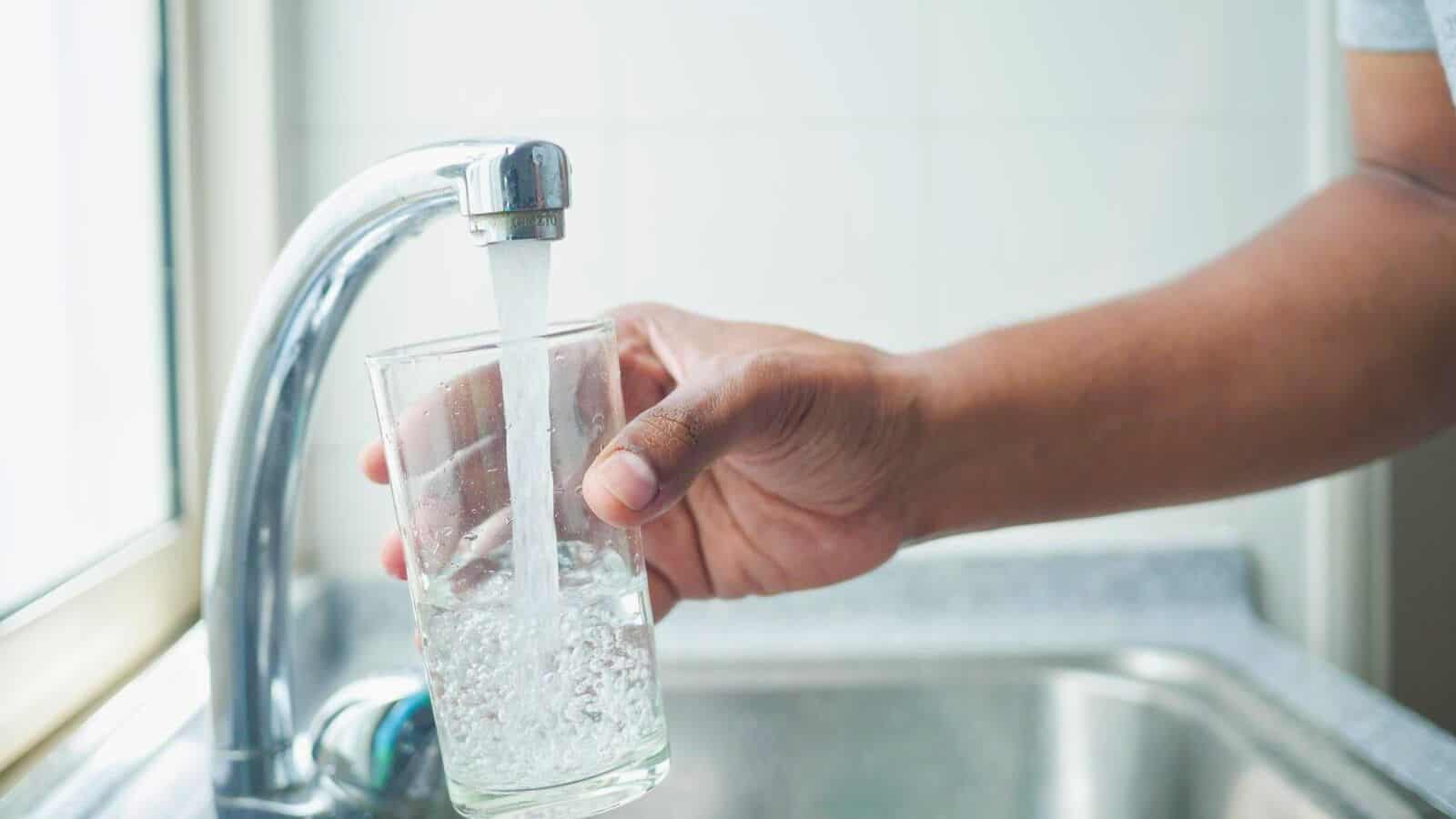 A person fills a glass with water from a kitchen faucet over a sink. The water flows steadily into the clear glass, and the counter is visible beneath the tap. The background shows a tiled wall.