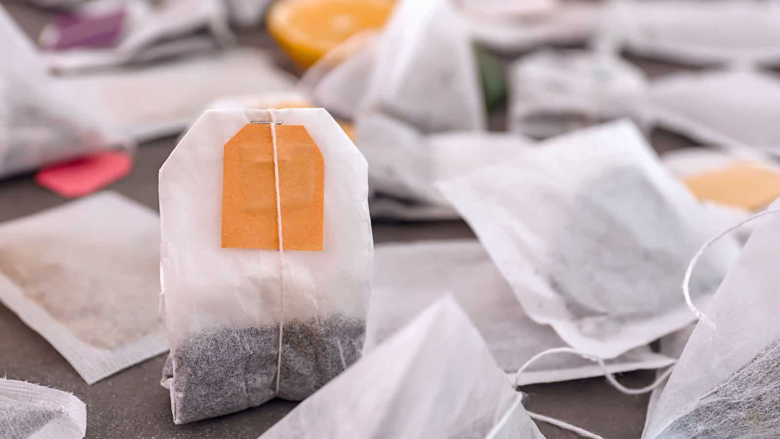 A collection of various tea bags is scattered on a surface. One upright tea bag with an orange tag is prominently in the foreground, while others lie flat in the background, featuring tags in different colors.