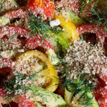A colorful and easy salad features sliced tomatoes and avocados, garnished with fresh dill and a crumbly topping, all spread on a white plate to brighten up your meal.