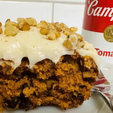 A slice of carrot cake topped with cream cheese frosting and walnuts sits on a plate with a fork, showcasing one of those timeless recipes. Nearby, a can of Campbell's Tomato Soup complements this raving delight.