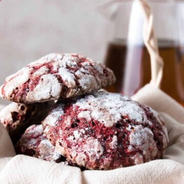 A basket containing three red crinkle cookies dusted with powdered sugar, placed on a beige cloth. In the background, there is a glass pitcher filled with a light brown liquid.
