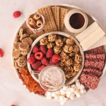 A round platter displaying assorted snacks including raspberries, granola clusters, breadsticks, wafers, rolled cookies, rice cakes, and dip. Additional raspberries are scattered on the marble surface around the platter.