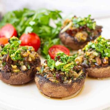Four stuffed mushrooms on a white plate, filled with a mixture of chopped herbs, nuts, and seasonings. There are fresh parsley and cherry tomatoes in the background.
