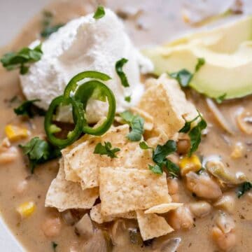 A bowl of creamy soup with corn, beans, and herbs, topped with tortilla chips, sliced jalapeños, avocado, and a dollop of sour cream. The dish is garnished with fresh cilantro.