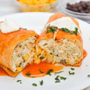 A plate with two pieces of a stuffed burrito, topped with red sauce and sour cream. The filling includes shredded chicken, rice, and corn. Garnished with chopped herbs. Background shows bowls of diced tomatoes and black beans.
