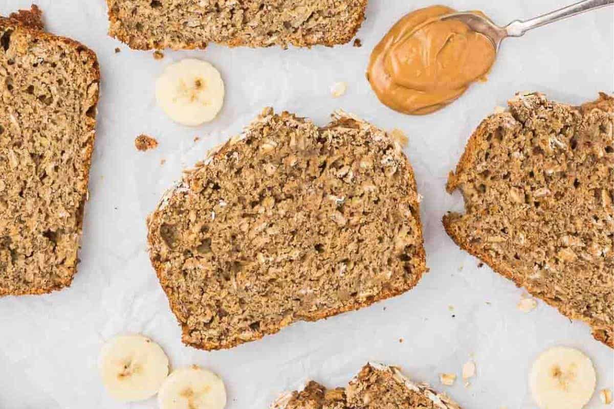 Slices of delicious banana bread rest on a white surface, surrounded by fresh banana slices and a spoonful of peanut butter—perfect for those who cherish bakery-inspired treats.