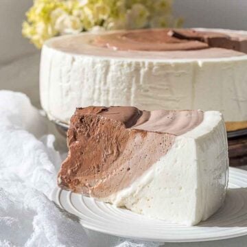 A slice of mousse cake on a white plate, featuring layers of chocolate and vanilla. The whole cake is in the background, partially cut. A jar of whipped cream and yellow flowers are also visible on the table.