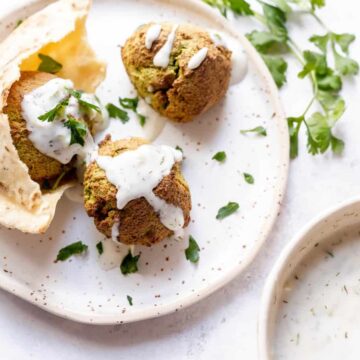 A plate with three falafel balls drizzled with white sauce, garnished with parsley, and served with pita bread—a feast of Middle Eastern flavors ready to transport your taste buds. A bowl of the same delicious sauce is nearby on a light-colored surface.