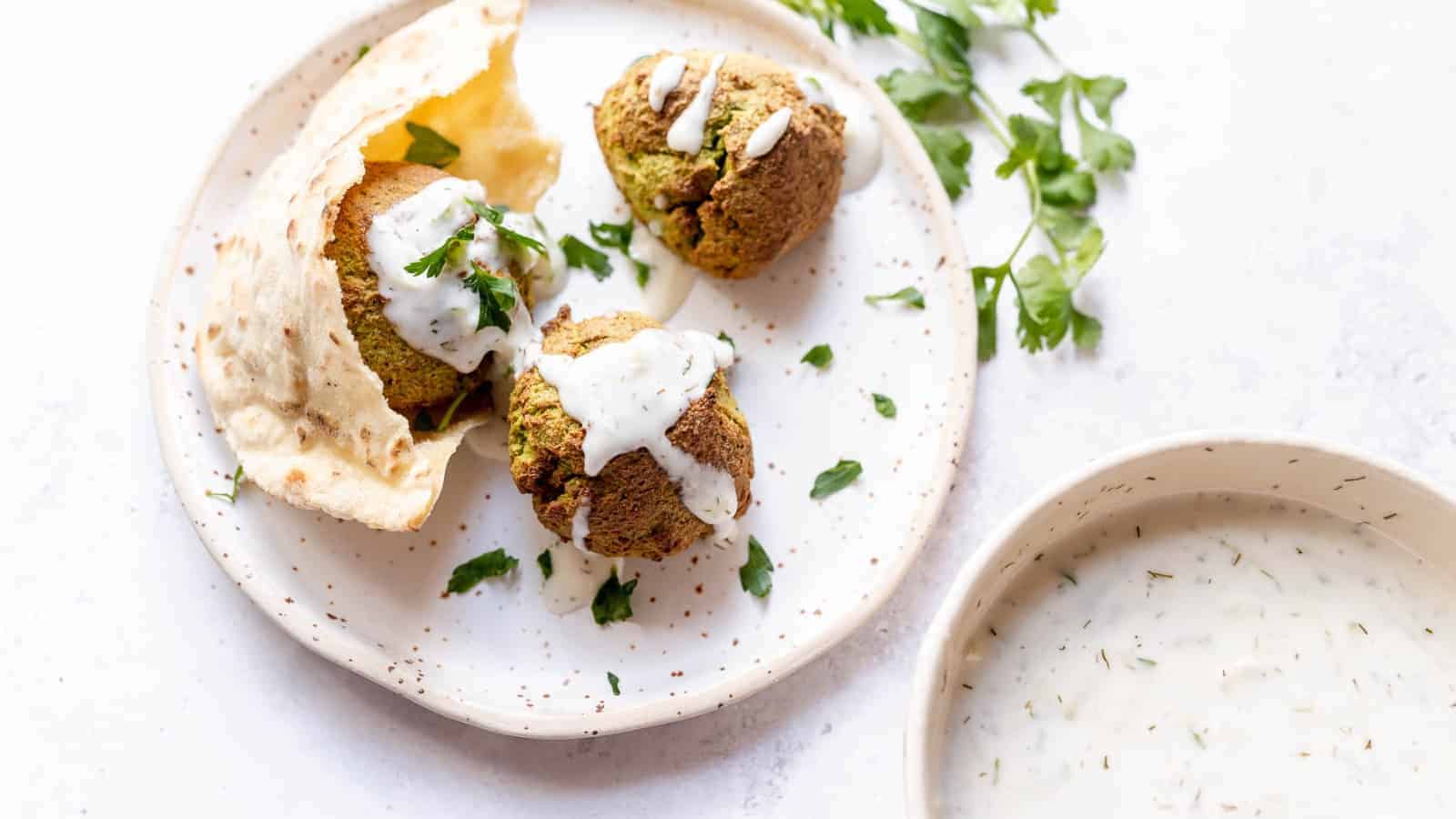 A plate with three falafel balls drizzled with white sauce, garnished with parsley, and served with pita bread—a feast of Middle Eastern flavors ready to transport your taste buds. A bowl of the same delicious sauce is nearby on a light-colored surface.