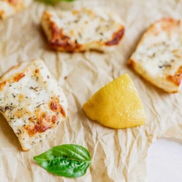 Grilled cheese pieces and lemon wedges are arranged on crumpled parchment paper. Fresh green basil leaves are scattered around the food.