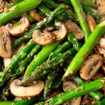 A plate of sautéed asparagus and sliced mushrooms seasoned with sesame seeds and black pepper. The vegetables are glossy, indicating they are cooked, and are arranged in a vibrant, fresh display.