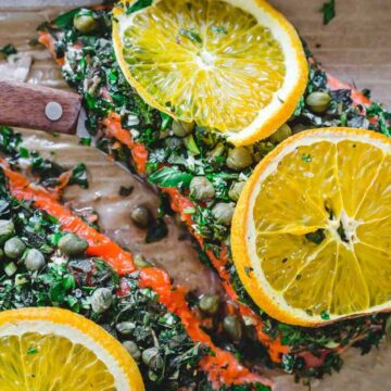 A delicious cooked salmon fillet is adorned with chopped herbs, capers, and vibrant orange slices on a parchment-lined surface. A wooden handle peeks into view, hinting at a knife ready to serve the delectable fish.
