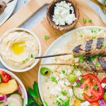 A plate with grilled meat skewers on rice, served with sliced tomatoes, onions, olives, and feta cheese. Adjacent bowls contain hummus with olive oil, a fresh salad with cucumbers and tomatoes, and a separate bowl of olives. A wooden pepper mill is nearby.