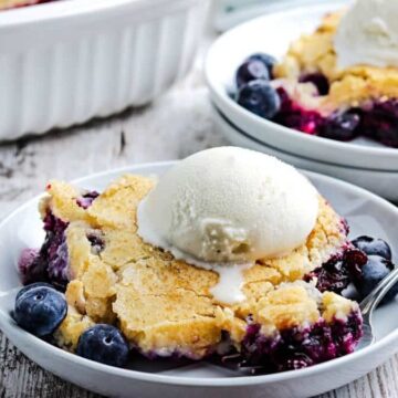 A slice of blueberry cobbler topped with a scoop of vanilla ice cream is served on a white plate, reminiscent of Grandma's dishes. Fresh blueberries are scattered around, tempting one to fight over the last bite. A spoon rests on the plate, with another serving and a baking dish in the background.