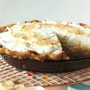 A pie with a golden brown crust and fluffy meringue topping is placed on a round wire rack, inviting you into the world of desserts. A slice is missing, revealing its creamy filling. The background includes a colorful cloth and part of a floral pattern, reminiscent of classic pie recipes.