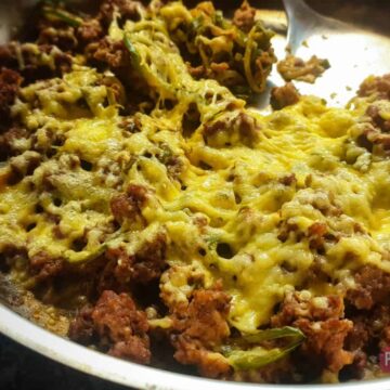 A skillet filled with a cooked dish featuring ground meat, melted cheese, and bits of green vegetables offers a perfect budget-friendly dinner idea. A spoon is partially visible in the upper right corner of the skillet.