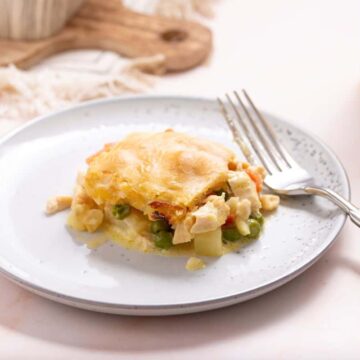 A portion of chicken pot pie, a classic from old-school recipes, is served on a white plate with a fork beside it. The pie boasts a golden, flaky crust and visible pieces of chicken, peas, and carrots. A glass of orange juice is partially seen on the right, ready to complete this family table favorite.