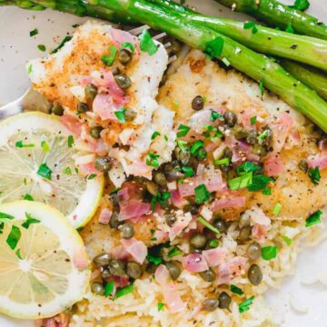 A plate of fish topped with diced onions and capers, served with slices of lemon. The fish is accompanied by a serving of rice and several asparagus spears. A fork is placed on the side of the plate.