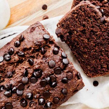 A delicious bread loaf speckled with chocolate chips sits on a light cloth, showcasing two neatly cut slices beside it. Nearby, a bottle of milk rests on the wooden surface, complementing this delectable creation straight from a beloved bakery's repertoire.
