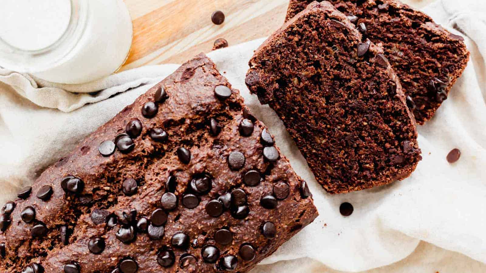 A delicious bread loaf speckled with chocolate chips sits on a light cloth, showcasing two neatly cut slices beside it. Nearby, a bottle of milk rests on the wooden surface, complementing this delectable creation straight from a beloved bakery's repertoire.