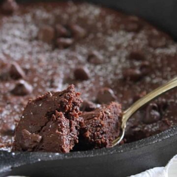 A close-up of a skillet brownie with chocolate chips sprinkled on top. A piece has been scooped, revealing the moist texture inside. A silver spoon with a piece of brownie rests on the edge of the skillet.