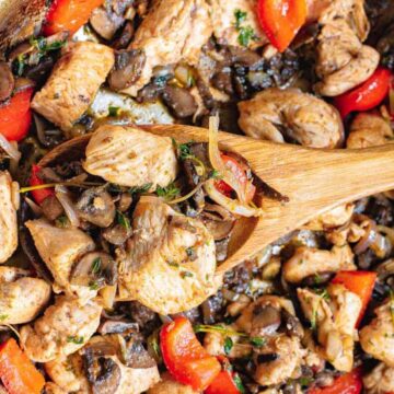 A skillet brimming with cooked chicken pieces, vibrant red bell peppers, sliced mushrooms, and onions. A wooden spoon lifts the savory mix, revealing its delicious texture. Perfect for when you're too tired to fuss over dinner—just wing it and enjoy this herb-garnished delight!.