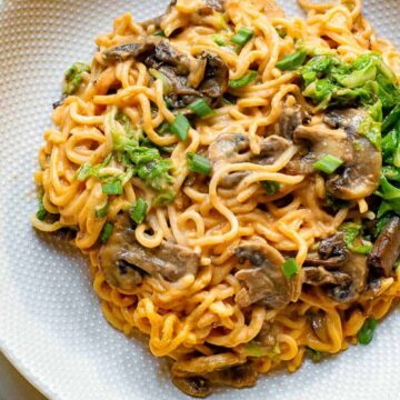 A white plate of creamy pasta topped with sliced mushrooms and green onions. The dish is garnished with additional green herbs. A napkin and chopsticks are partially visible in the background.