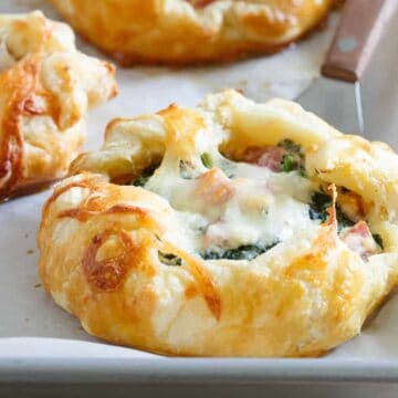 A baking tray with pastries topped with melted cheese, spinach, and diced ham. The pastries are golden brown with flaky, crusty edges. A knife is partially visible on the tray lined with parchment paper.