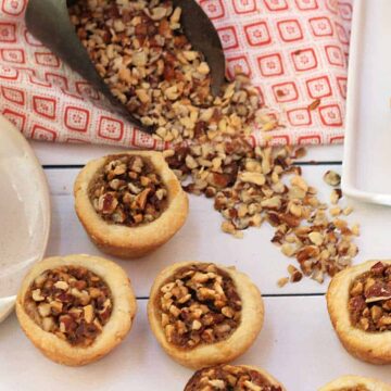 A cup of tea sits on a saucer next to homemade pastries topped with chopped nuts, reminiscent of old-fashioned meals. Additional nuts are scattered beside a metal scoop on a red and white patterned cloth, evoking warm memories amid family feuds. Some pastries are arranged on a white platter.