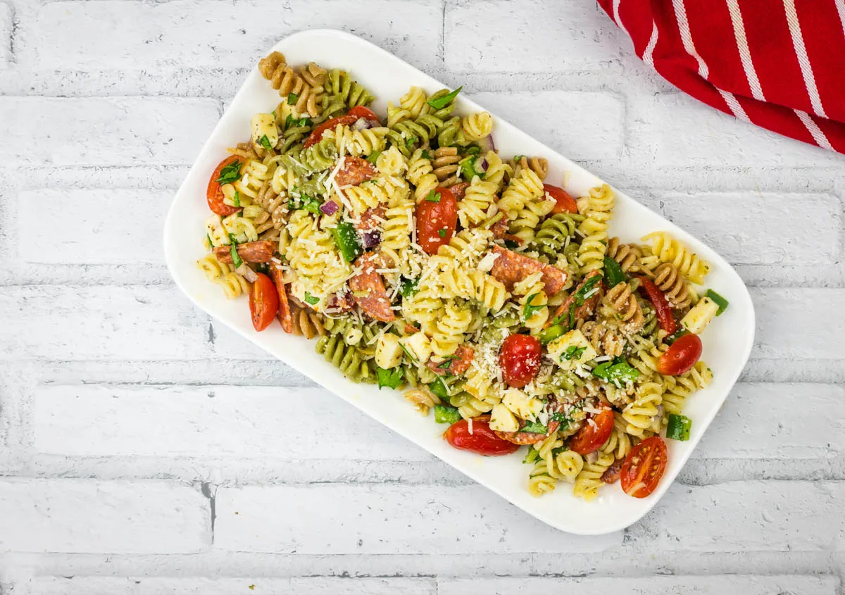 A rectangular white plate holds a vibrant pasta salad with corkscrew-shaped noodles, cherry tomatoes, cubed cheese, and assorted greens—perfect for healthy recipes. A red and white striped cloth is partially visible on the smooth white brick surface.