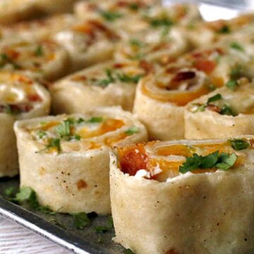 A tray of rolled tortillas filled with cheese, diced vegetables, and herbs. The rolls are sliced into pinwheels and garnished with chopped cilantro.