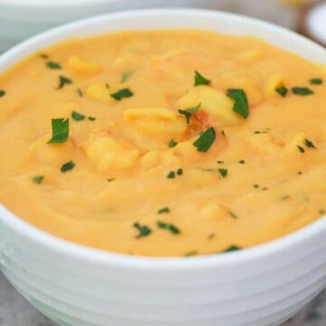 A white bowl filled with creamy orange soup, garnished with chopped green herbs. A black spoon and striped cloth are nearby on a table.