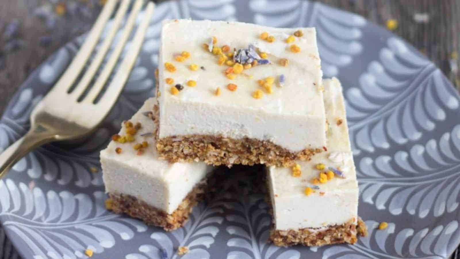 A plate with three stacked pieces of creamy dessert bars, topped with small yellow and purple sprinkles. The bars have a crumbly brown base. A fork is placed next to the bars on the patterned plate.