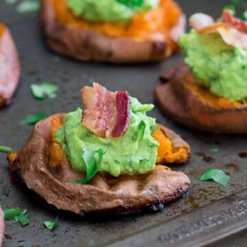 Baked sweet potato slices topped with mashed avocado and bacon pieces are arranged on a dark baking sheet. Fresh herbs are sprinkled on top. The dish is presented on a wooden surface.