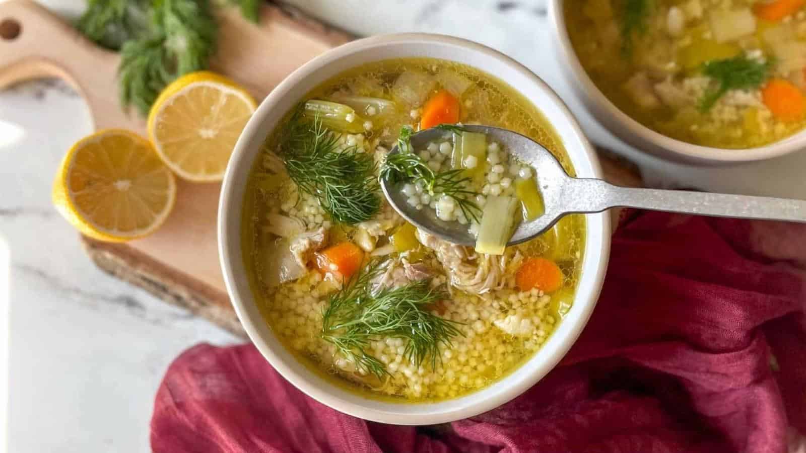 A bowl of soup containing small pasta, chunks of vegetables, and fresh dill, with a spoon lifting some soup. A wooden board with lemon slices and dill is in the background, alongside a red cloth.