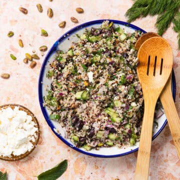 A vibrant quinoa cucumber salad with olives, red onions, and herbs sits on a marble surface. Wooden salad servers rest on the bowl's edge. Nearby, small bowls hold feta cheese and pistachios. Sprigs of dill and mint leaves are artfully scattered around.