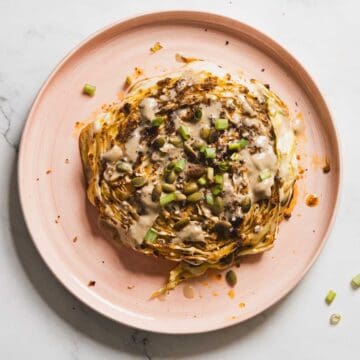 A pink plate hosts a large slice of grilled cabbage, charred to perfection, drizzled with creamy tahini sauce. Garnished with chopped green onions and seeds, these air fryer cabbage steaks rest elegantly on a white marble surface.