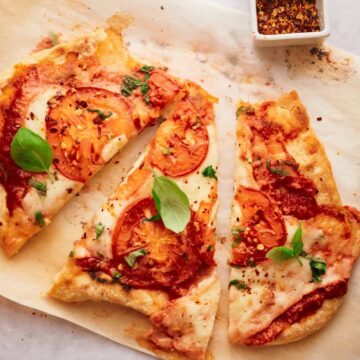 A sliced pizza rests on parchment paper, topped with tomato slices, melted cheese, fresh basil, and red pepper flakes. A small container of additional red pepper flakes is nearby on the white surface.