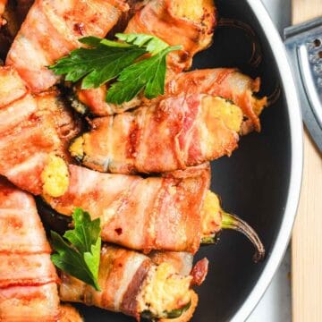 A dish of bacon-wrapped jalapeño poppers is arranged in a pan, garnished with parsley. Grated cheese and a grater are on a wooden cutting board nearby. The poppers are filled with a creamy cheese mixture and have a golden-brown, crispy appearance.